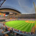 Hands holding a digital trading device above a crowd in a baseball stadium at night.