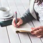 Hands holding a smartphone showing audio waveform on a bright desk with a mug and notepad