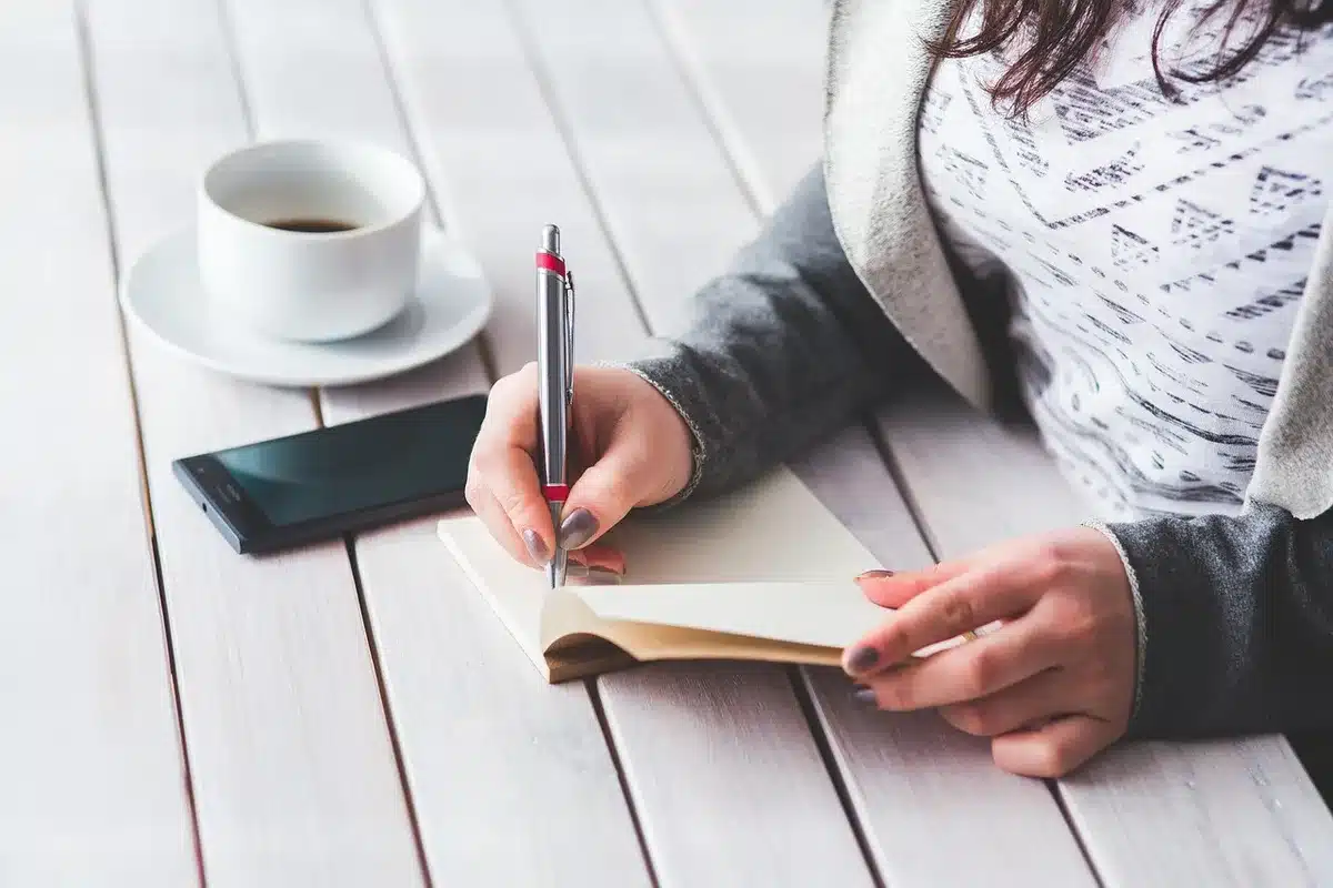 Hands holding a smartphone showing audio waveform on a bright desk with a mug and notepad