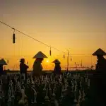 Three silhouetted figures walking past a small wooden church near a riverbank at dusk
